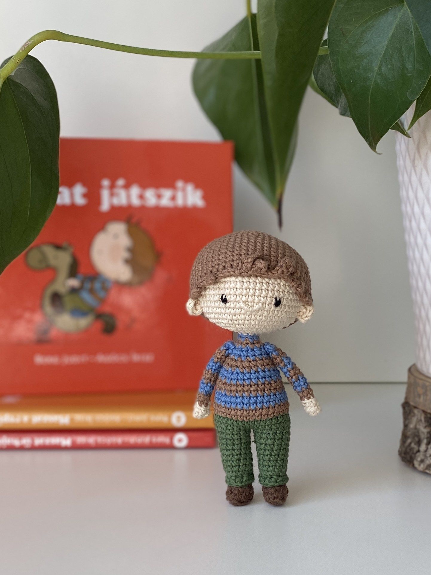 Kézzel horgolt Maszat figura kék-barna csíkos pulóverben és zöld nadrágban, mögötte egy Maszat-könyv